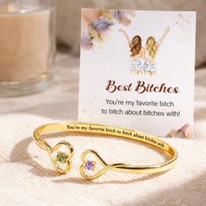 May include: A gold-colored open cuff bracelet with heart-shaped ends, each holding a gemstone. One gemstone is green, the other is purple. A card behind the bracelet reads "Best Bitches" and a quote.