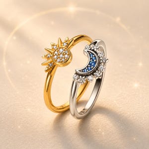 Può includere: Sono mostrati due anelli, uno dorato e uno argentato. L'anello dorato presenta un design a sole con un cerchio di piccole pietre trasparenti e raggi appuntiti. L'anello argentato ha una luna crescente con pietre blu e dettagli a stella. Entrambi gli anelli hanno una finitura liscia e lucida.