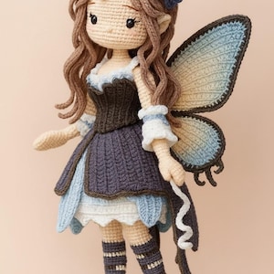 Peut inclure: Une poupée fée faite à la main au crochet, avec des cheveux bruns, des ailes bleues et blanches et un ornement floral. La poupée porte une robe bleu foncé et marron avec une bordure blanche et des chaussettes rayées. La poupée fée mesure environ 25 cm de haut.