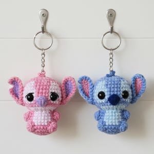 May include: Two crochet keychains, one pink and white, the other blue and light blue, each featuring a cartoon character design. The keychains are hanging from silver metal key rings and chains. The keychains have large black eyes and large ears.