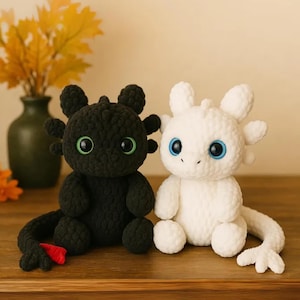 May include: Two crocheted dragon plush toys. One is black with green eyes and a red fin, the other is white with blue eyes. Both have rounded features and are sitting on a wooden surface.