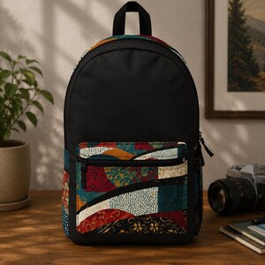 May include: A black backpack featuring a front pocket with a multi-colored abstract pattern. The pattern includes curved shapes in shades of red, teal, and cream with intricate dotted and floral textures. The bag has a top handle and black zippers.