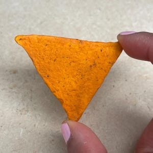 May include: A single, orange-colored tortilla chip in a triangular shape. The chip has a textured surface and is held between two fingers with pink nail polish.