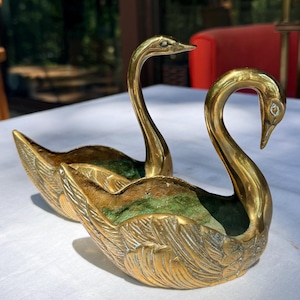 Puede incluir: Dos cuencos de latón pulido con forma de cisne, con texturas de plumas detalladas en las alas exteriores y un acabado de pátina verde en el interior. Las figuras de cisne tienen cuellos largos y curvados con las cabezas orientadas hacia abajo.