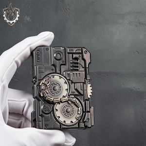 Op de afbeelding: Een Zippo aansteker met een gedetailleerd, industrieel ontwerp. De metallic grijze aansteker heeft tandwielen, klinknagels en een kettingachtig element. Het woord "Zippo" is zichtbaar aan de zijkant. Het ontwerp heeft een steampunk esthetiek.