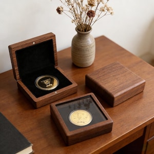Op de afbeelding: Een gouden munt gepresenteerd in een houten kistje met een zwarte fluwelen binnenkant. De munt wordt ook getoond in een vierkante houten vitrine met een doorzichtig deksel. Een gesloten houten kistje en een kleine vaas met droogbloemen zijn ook aanwezig.