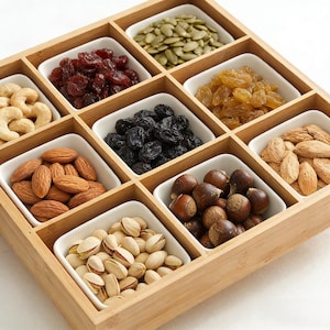 Könnte beinhalten: Ein Holzserviertablett mit neun weißen quadratischen Schalen, gefüllt mit verschiedenen Nüssen und Trockenfrüchten. Das Tablett enthält Cashewnüsse, Mandeln, Pistazien, Haselnüsse, Rosinen, Cranberries und Kürbiskerne.