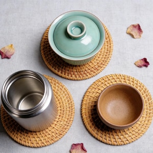 Könnte beinhalten: Draufsicht auf drei runde Untersetzer aus Weide, die jeweils einen anderen Gegenstand tragen. Ein hellgrünes Keramik-Teeservice mit Deckel, ein Edelstahlbehälter und eine kleine braune Keramikschale sind zu sehen. Verstreute Rosenblätter setzen Farbakzente.