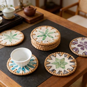 Könnte beinhalten: Eine Sammlung runder Untersetzer mit einem gewebten Rattanrand. Jeder Untersetzer hat ein einzigartiges Blumendesign in Grün-, Blau- und Lilatönen. Eine weiße Teetasse steht auf einem Untersetzer mit blauem Blättermuster. Die Untersetzer sind auf einer dunklen Oberfläche angeordnet.