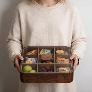 Könnte beinhalten: Eine Snackbox aus Holz mit einem transparenten Deckel, unterteilt in neun Fächer. Die Box ist mit verschiedenen Snacks gefüllt, darunter verpackte Leckereien, Früchte und Nüsse. Die Box wird von einer Person gehalten, die einen beigen Pullover trägt.