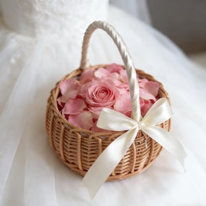 Könnte beinhalten: Ein runder Weidenkorb gefüllt mit rosa Rosenblättern und einer einzelnen rosa Rose. Der Korb hat eine weiße Satin-Schleife und einen passenden Griff. Der Korb ist wahrscheinlich für eine Hochzeit.