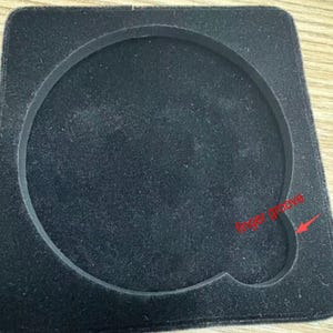 Puede incluir: Inserto negro de forma cuadrada con una hendidura circular y una ranura para los dedos. El inserto est&aacute; hecho de un material suave y aterciopelado, y la hendidura circular est&aacute; delineada con un borde elevado.