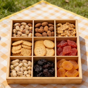 Könnte beinhalten: Ein Holztablett mit neun Fächern, die jeweils mit verschiedenen Snacks gefüllt sind. Das Tablett enthält Pistazien, Mandeln, Sesamkrokant, Cracker, getrocknete Papaya, Rosinen und getrocknete Aprikosen. Das Tablett steht auf einer gelb-weiß karierten Picknickdecke.