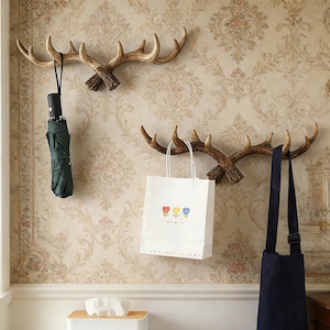 Könnte beinhalten: Zwei geweihförmige Wandhaken aus braunem Material sind an einer gemusterten Wand befestigt. Ein Haken hält einen grünen Regenschirm, der andere eine weiße Papiertüte mit Blumendesigns und eine marineblaue Tasche.