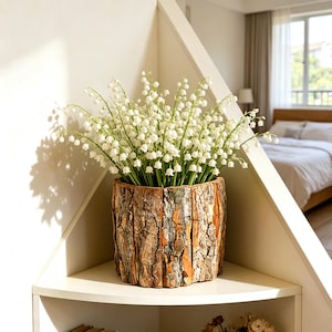 Könnte beinhalten: Ein Arrangement aus weißen Maiglöckchen in einem rustikalen Pflanzgefäß mit Rindenstruktur. Der braune Pflanztopf mit orangefarbenen Akzenten steht auf einem weißen Regal. Die Blumen haben kleine, glockenförmige weiße Blüten und grüne Stiele.