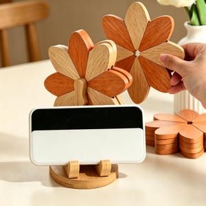 May include: Wooden flower-shaped trivets in shades of orange and natural wood. A white and black smartphone rests on a wooden stand. The trivets are designed to resemble flower petals and are likely used for hot dishes.