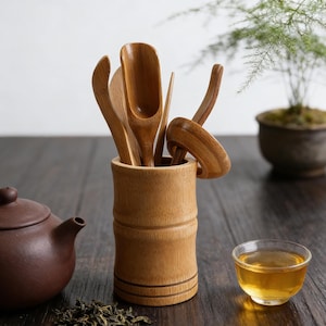 Könnte beinhalten: Ein Teeset mit einer braunen Teekanne, einem Bambusbehälter mit hölzernen Teeutensilien und einer kleinen gläsernen Teetasse mit Tee. Getrocknete Teeblätter liegen auf der dunklen Holzoberfläche.