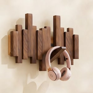 May include: A wooden wall-mounted coat rack with a series of vertical, rectangular wooden pieces in varying heights and widths. A pair of pink headphones hangs from the rack. The rack is made of dark brown wood.