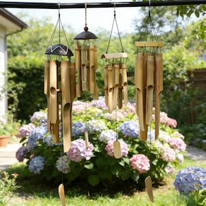 May include: Three bamboo wind chimes hanging outdoors. Each chime features natural bamboo tubes and wooden accents. The chimes are suspended from a black metal bar, with a backdrop of lush green foliage and colorful hydrangeas.