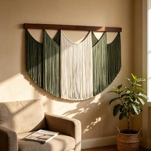 Può includere: Un arazzo da parete con lunghi cordoni verticali in cotone sospesi a una barra orizzontale in legno scuro. Il design presenta sezioni alternate di cordoni color verde oliva e crema disposti in una serie di curve drappeggiate e festonate.