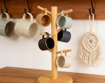 May include: A wooden mug tree with a round base and multiple branches holding various ceramic mugs. The mugs are in shades of beige, blue, and black. A macrame wall hanging is visible on the right, and other mugs hang on a wooden shelf.