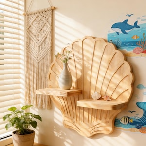 Könnte beinhalten: Ein wandregal aus Holz in Muschelform mit zwei Ablagen. Eine kleine Vase mit Blumen und zwei Muscheln befinden sich auf den Regalen. Ein Makramee-Wandbehang und eine Topfpflanze sind ebenfalls zu sehen.