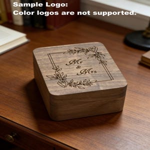 Puede incluir: Una caja de madera cuadrada con tapa con bisagras. La tapa est&aacute; grabada con las palabras "Mr & Mrs" dentro de un marco rectangular, rodeado de un dise&ntilde;o floral. La caja es de madera marr&oacute;n claro y se encuentra sobre una superficie marr&oacute;n oscuro.