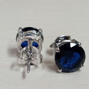 May include: A pair of stud earrings featuring round, deep blue faceted stones set in a silver-toned metal prong setting. One earring is shown from the front, while the other is positioned to display the back post and the secure metal butterfly clutch.