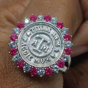 May include: A silver ring with a circular emblem inscribed with "TEXAS A&M AGGIE MOM" and "A M". The ring is embellished with alternating pink and blue gemstones encircling the central design.