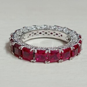May include: A silver-toned eternity band ring featuring a continuous row of square-cut, deep red stones. The metal setting is detailed with small, clear pavé-set stones along the edges, creating a textured, sparkling border around the red gems.