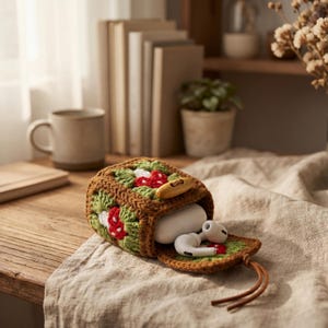 May include: A brown, green, and red crocheted case with a wooden button closure, holding white earbuds. The case features a mushroom design and a leather drawstring. The case is resting on a beige cloth on a wooden surface.