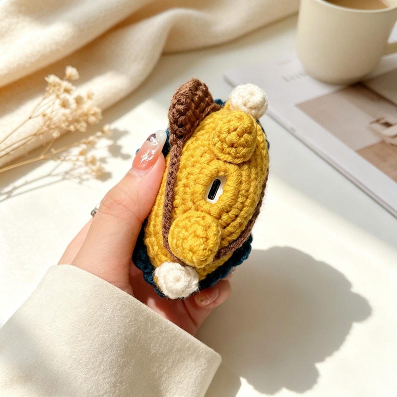 May include: A crochet earbud case cover featuring a yellow body with a dark brown trim and small white circular accents. The case has a visible cutout for a charging port and a textured, handmade yarn construction with a soft, rounded shape.