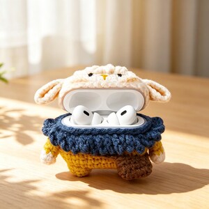 May include: A crochet case for wireless earbuds shaped like a sheep with a cream-colored head, a dark blue ruffled collar, a yellow body, and a small brown rectangular pouch attached to the side, holding white earbuds inside the open lid.