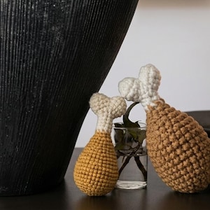 May include: Two handmade crochet chicken drumsticks, one mustard yellow and one brown, with white bone details. They are displayed near a black vase, a lit candle, and a small glass of greenery. A decorative item for the home.