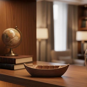 Puede incluir: Un cuenco decorativo de madera con forma de barco está sobre una estantería de madera marrón oscuro. Detrás, un globo terráqueo de latón y dos libros. El fondo muestra una habitación con lámparas y cortinas. El cuenco mide unos 30 cm de largo.