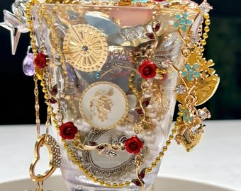 May include: A clear glass goblet filled with an assortment of jewelry and decorative items. Gold chains, red rose accents, and a heart-shaped pendant with a turquoise center are visible. The goblet is filled with pearls and other trinkets.