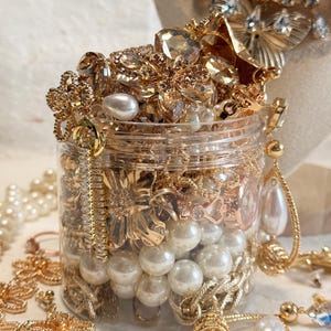 Puede incluir: Un frasco de plástico transparente lleno de joyas doradas, incluyendo varios collares, cadenas y cuentas de perlas blancas artificiales. El frasco está rebosante de colgantes metálicos en forma de flor, cristales claros y pendientes colgantes.