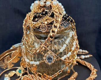 May include: A collection of gold-toned jewelry, including necklaces, bracelets, and earrings, spilling from a decorative glass jar. The jewelry features pearls, gemstones, and various chain styles. The jar sits on a dark surface.