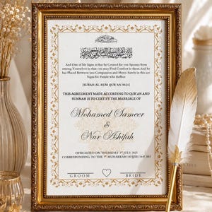 May include: A rectangular gold-toned ornate frame containing a white marriage certificate with black Arabic and English text, including the names Mohamed Sameer and Nur Ashifah, featuring a decorative gold border and a white and gold feather quill pen.