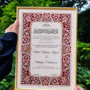 May include: A rectangular marriage certificate in a gold frame featuring a deep red border with intricate floral patterns. The text includes Quranic verses, the names Abul Kalam Azad and Shifaya Nashreen, and the date 14th July 2024 in black script.