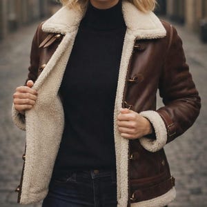 Puede incluir: Chaqueta de cuero marr&oacute;n con forro y cuello de borrego color crema. La chaqueta tiene cierres de palanca y detalles de cintur&oacute;n con hebilla en los pu&ntilde;os. Se lleva un cuello alto negro y vaqueros azul oscuro.