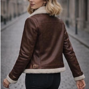 Puede incluir: Chaqueta de cuero marr&oacute;n con cuello y forro de borreguillo color crema. La chaqueta tiene un estilo aviador cl&aacute;sico con cintura y pu&ntilde;os abrochados. La modelo lleva vaqueros negros.