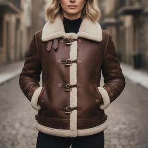 Puede incluir: Chaqueta de cuero marr&oacute;n con cuello y forro de borrego color crema. La chaqueta presenta cierres de palanca y ribete de borrego en los pu&ntilde;os y bolsillos. Una prenda de abrigo elegante.