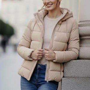 Puede incluir: Chaqueta acolchada beige con capucha, cierre de botones y cremallera. La chaqueta se lleva sobre un jersey de color claro y se combina con vaqueros azules. La chaqueta tiene un diseño acolchado y es adecuada para el frío.