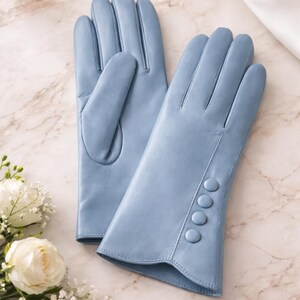Peut inclure: Une paire de gants en cuir bleu clair. Les gants ont un design classique avec quatre boutons décoratifs sur le poignet. Ils sont présentés sur une surface en marbre, avec un flacon de parfum, des perles et des fleurs blanches en arrière-plan.