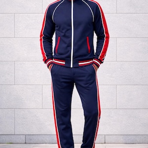 Op de afbeelding: Marineblauw trainingspak met rode en witte accenten. De jas heeft een witte rits en rode strepen op de mouwen. De bijpassende broek heeft een rode streep aan de zijkant. Witte sneakers.