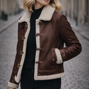 Puede incluir: Chaqueta de cuero marr&oacute;n con forro y ribete de borrego color crema. La chaqueta presenta un cuello con solapas, cierres de palanca y dos bolsillos delanteros. La chaqueta se lleva sobre un cuello alto negro.