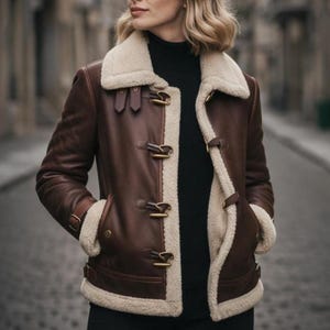 Puede incluir: Chaqueta de cuero marr&oacute;n con forro y cuello de borreguillo color crema. La chaqueta presenta cierres de palanca y un estilo aviador cl&aacute;sico. La modelo lleva un cuello alto negro. La chaqueta tiene dos bolsillos delanteros.