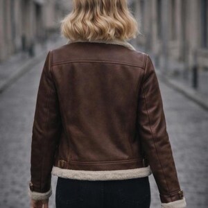 Puede incluir: Chaqueta de cuero marr&oacute;n con forro de borreguillo color crema en el cuello, pu&ntilde;os y dobladillo. La chaqueta tiene un estilo aviador cl&aacute;sico con detalles de hebilla en la cintura y las mangas. La persona lleva vaqueros negros.