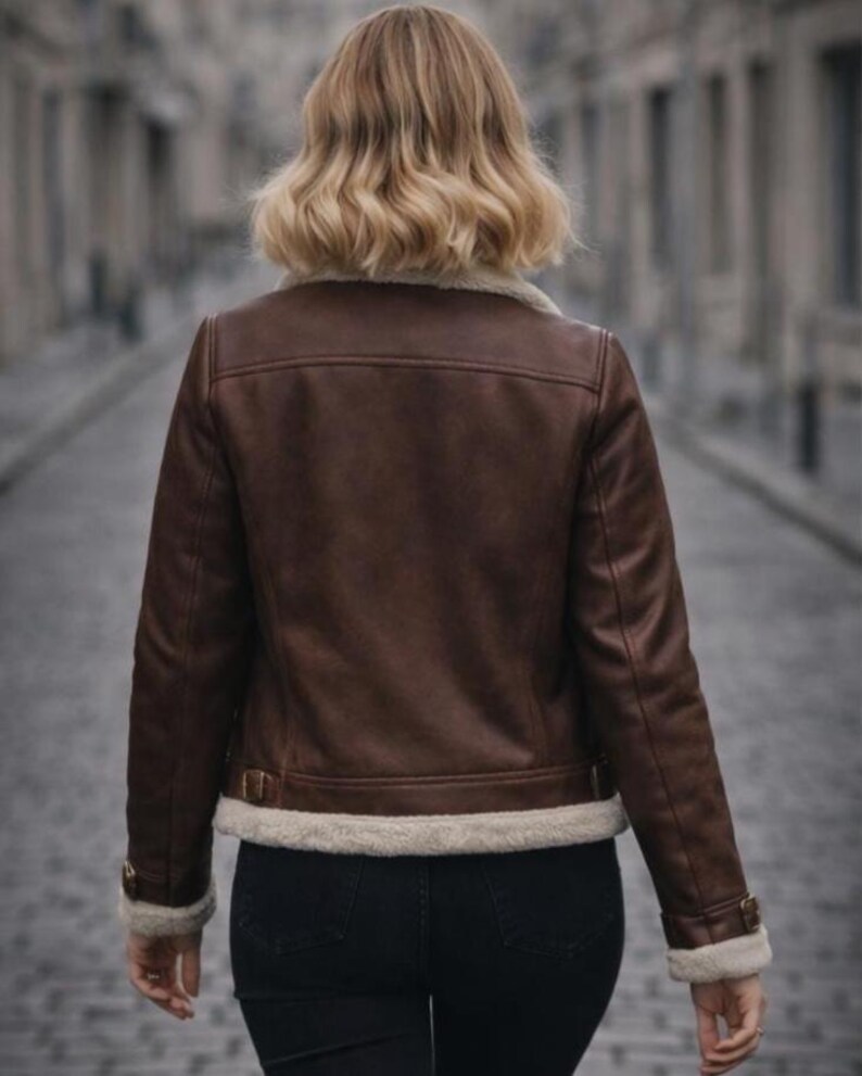 Puede incluir: Chaqueta de cuero marr&oacute;n con forro de borreguillo color crema en el cuello, pu&ntilde;os y dobladillo. La chaqueta tiene un estilo aviador cl&aacute;sico con detalles de hebilla en la cintura y las mangas. La persona lleva vaqueros negros.